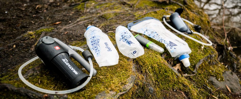 Selection of water filters on mossy background: 2 pumps, squeeze filter, UV purifier, and gravity filter